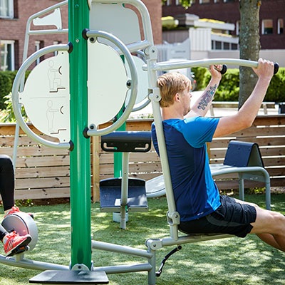 A man is working out on a multi gym unit which is outdoors in front of commercial buildings.